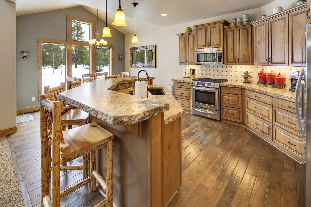 A sunny kitchen scene featuring a spacious island, natural wood cabinetry, high-end stainless steel appliances, and large windows that flood the area with daylight. The space feels modern yet grounded in nature, evoking the forest retreat ambiance described.