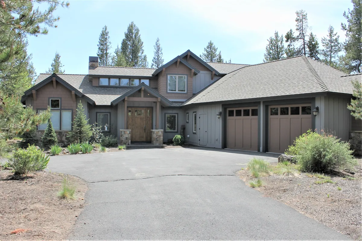 A welcoming view of the luxury home's front entrance framed by towering trees, with a spacious driveway and garage doors visible. The setting feels peaceful and nestled in nature, hinting at access to nearby walking trails and outdoor adventures just steps from the door.