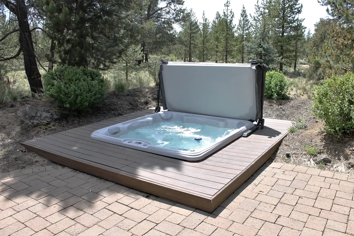 This image showcases the home's in-ground hot tub on a wooden deck surrounded by lush greenery and towering pines, creating a tranquil outdoor oasis. The peaceful, secluded setting is illuminated softly, ideal for stargazing or relaxing in the evening. The image suggests both privacy and a direct connection to the natural beauty of Central Oregon.