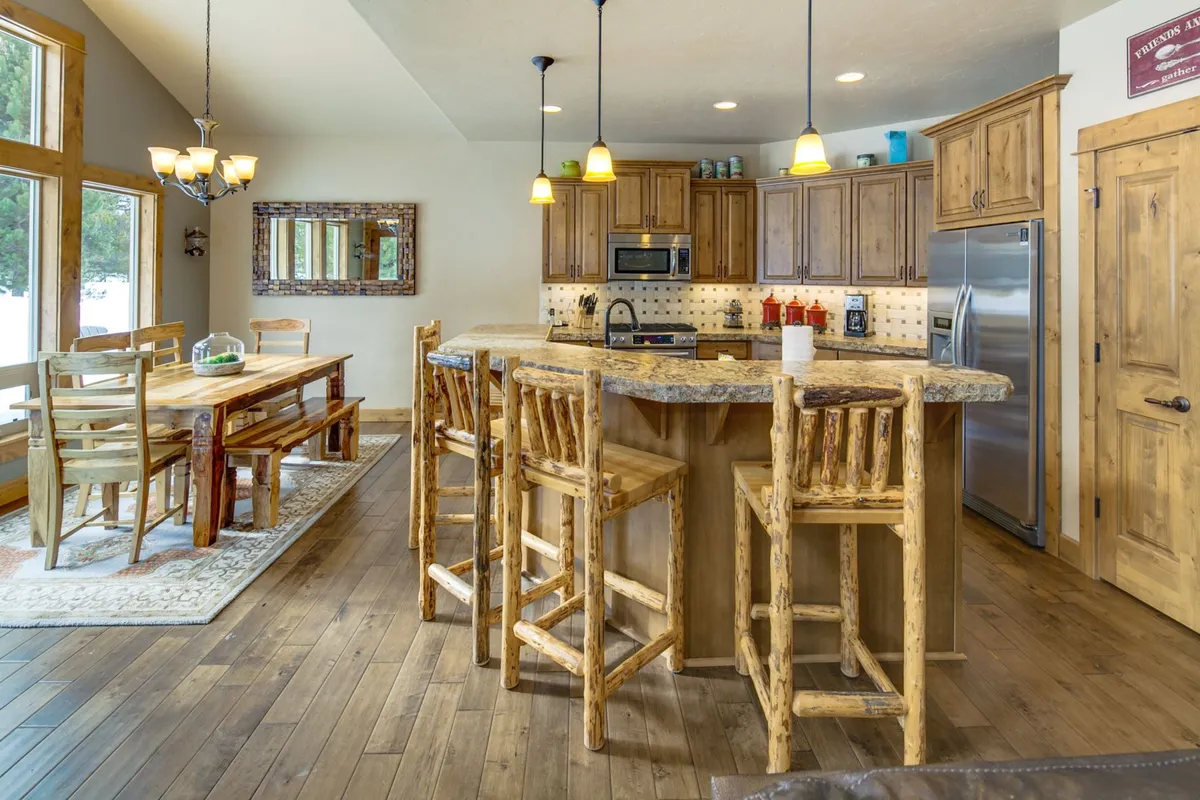 A wide shot of the open kitchen and dining area featuring a rustic kitchen island with bar stools, a grand dining table ready for large meals, expansive windows allowing in forested views, and warm wooden cabinetry tying the spaces together. The layout encourages flow between cooking, dining, and socializing.
