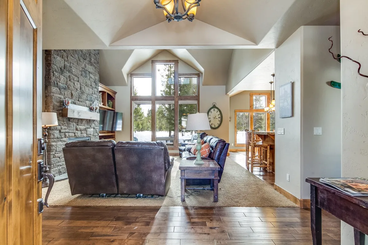This interior view features a spacious, inviting living area with a striking stone fireplace, large windows offering views of the outdoors, comfortable plush seating, and a dining area in the background. The room is styled for both relaxation and entertaining, blending luxury with a cozy, welcoming atmosphere.