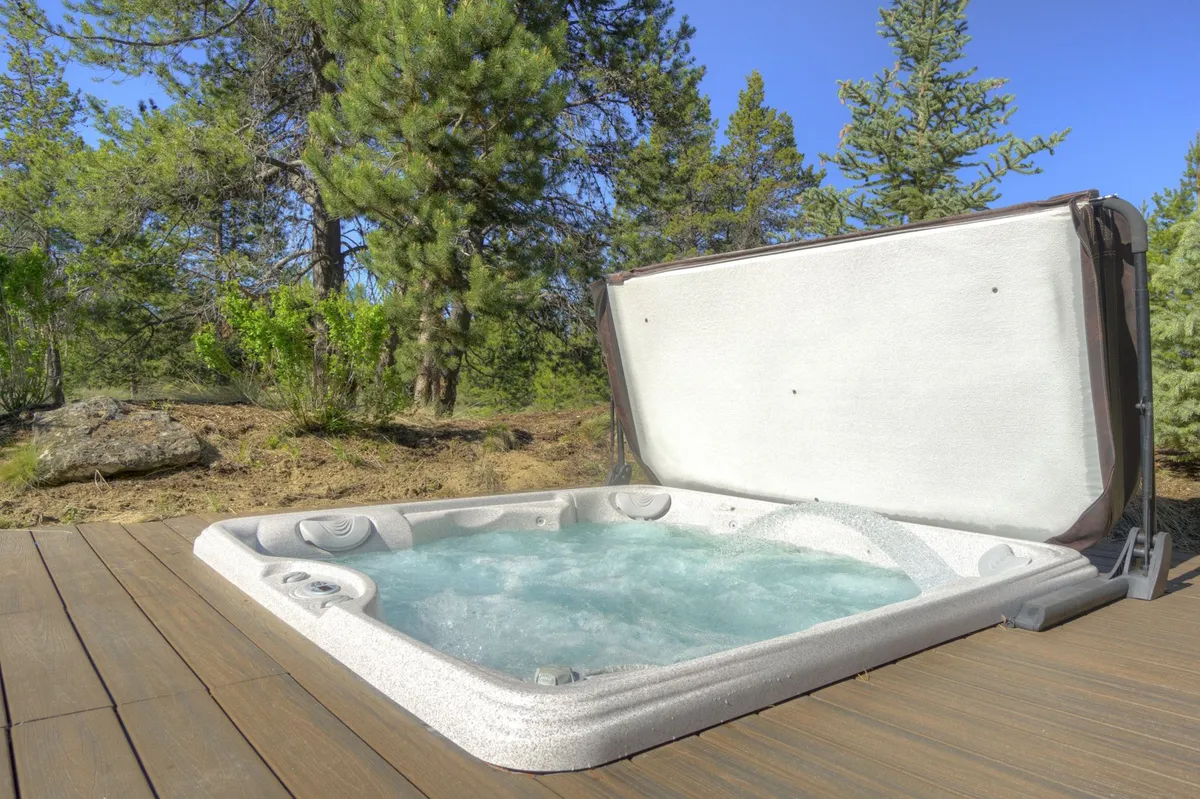 An inviting outdoor hot tub sits on a wooden deck surrounded by lush trees, providing a tranquil setting that blends seamlessly with nature. The expansive decking offers room for relaxation and socializing, with the forest backdrop hinting at the abundance of outdoor adventures available just steps away.
