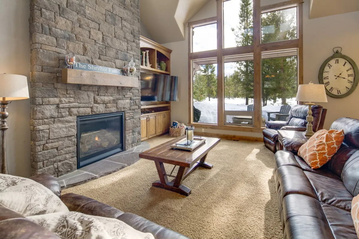 This image showcases a spacious living room with a prominent stone fireplace, large windows that invite natural light, a robust wooden coffee table, and comfortable leather sofas arranged for conversation and relaxation. The setting feels intimate and warm, perfect for family gatherings and unwinding after a long day.