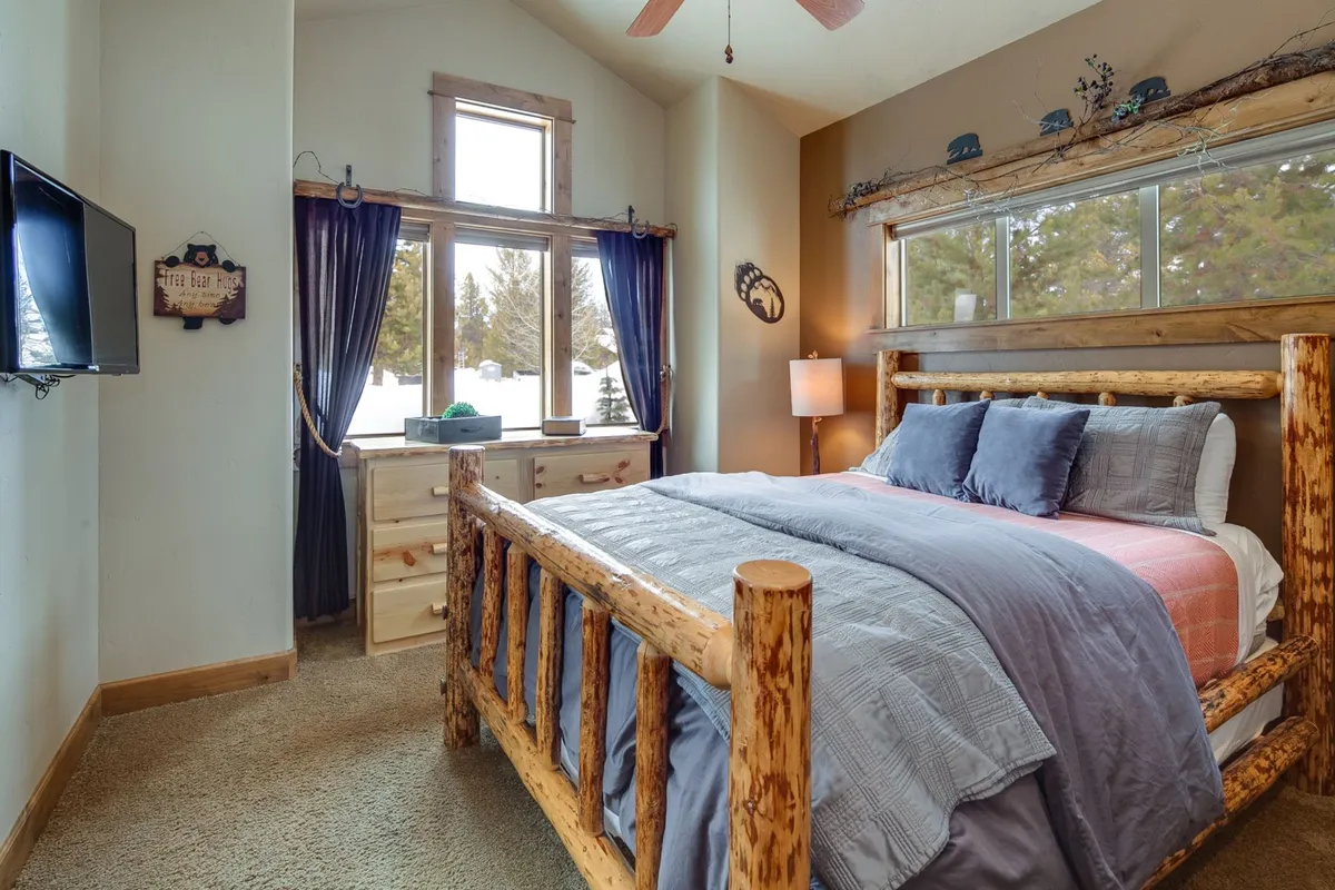 A spacious bedroom with a large log bed, soft bedding, and a wooden dresser. Light pours in through the large windows, revealing a tranquil view of the surrounding natural landscape. The overall ambiance is one of warmth, comfort, and relaxation, ideal for a primary bedroom retreat.