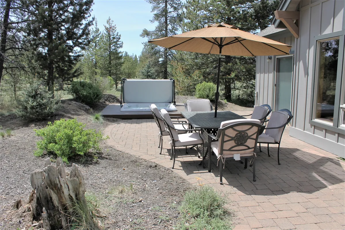 This exterior image showcases a spacious patio area with an outdoor dining table, umbrella, and a private hot tub set against a backdrop of lush trees. The setting invites guests to dine alfresco, soak in the hot tub, and enjoy the tranquility of nature.